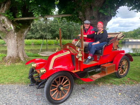 Club member Robert and TV personality Angel Strawbridge smiling in a red veteran Renault beside the water at Château de la Motte-Husson